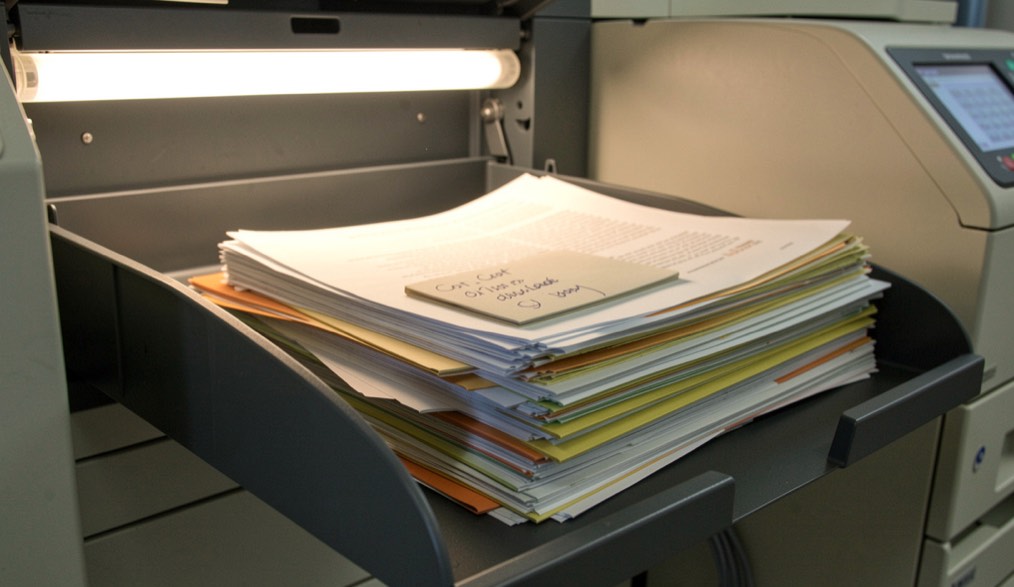 A photocopier tray holding a stack of printed pages beside a handwritten cost sheet